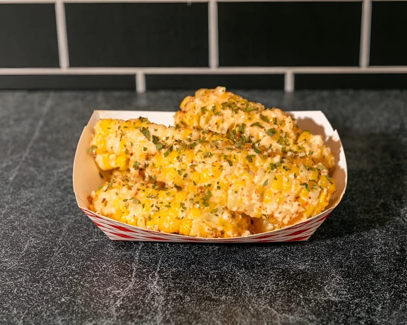 Elote-style corn ribs, coated in creamy sauce and sprinkled with herbs, served in a striped paper tray.