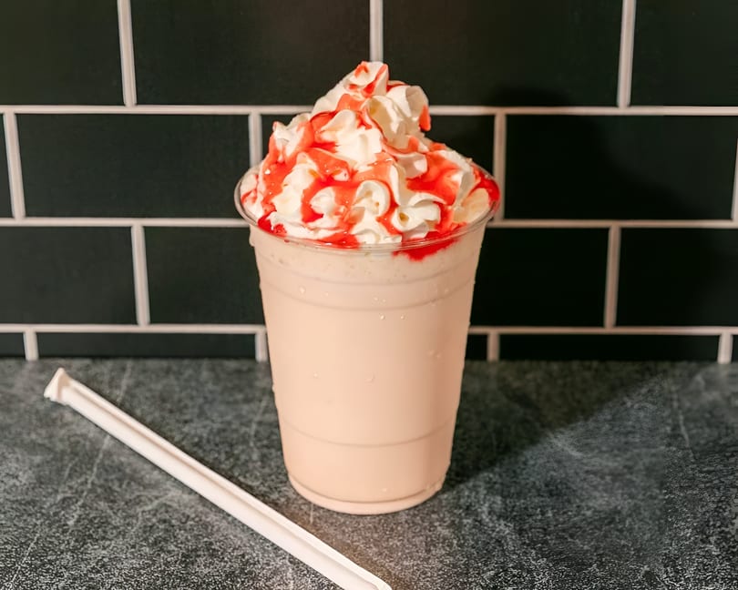 Strawberry milkshake with whipped cream and red syrup drizzle, next to a white straw.