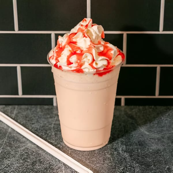 A strawberry milkshake with whipped cream and red syrup drizzle, next to a white straw.