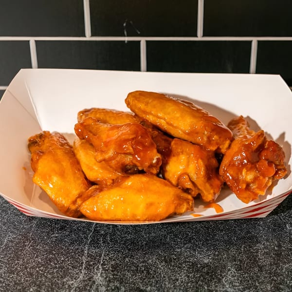 A white paper tray with red stripes holds a pile of saucy, orange-glazed chicken wings.