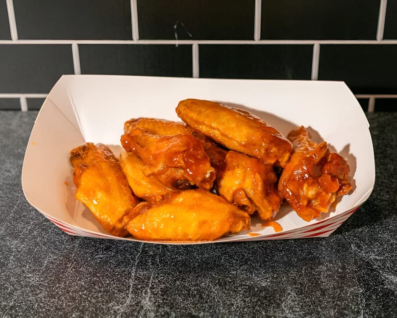 A white paper tray with red stripes holds a pile of saucy orange chicken wings.