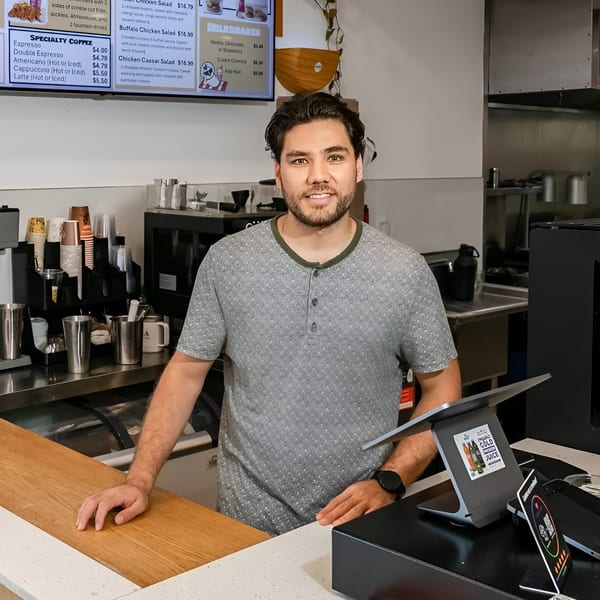 A smiling male barista stands behind a counter with coffee machines and a digital menu board.