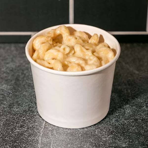 Creamy mac and cheese in a white to-go cup on a dark speckled counter with a black tiled wall.