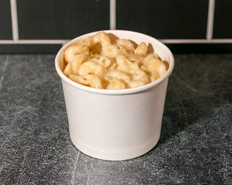 Creamy mac and cheese in a white to-go cup on a dark speckled counter with black tiles.